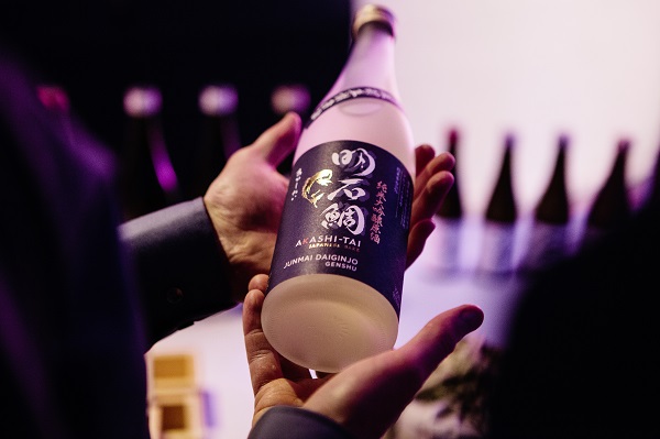 Eine Person hält eine Flasche Sake namens -Akashi-Tai- mit einem eleganten, farbigen Etikett. Im Hintergrund sind weitere Flaschen sichtbar, der gesamte Raum hat eine gedimmte Beleuchtung.