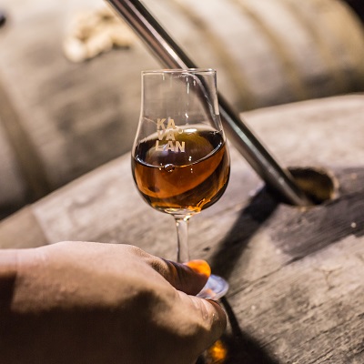 Ein Glas mit bernsteinfarbenem Whisky wird von einer Hand auf einem Holzfass gehalten. Im Hintergrund sind weitere Fässer zu sehen, was auf eine Brennerei-Umgebung hindeutet.