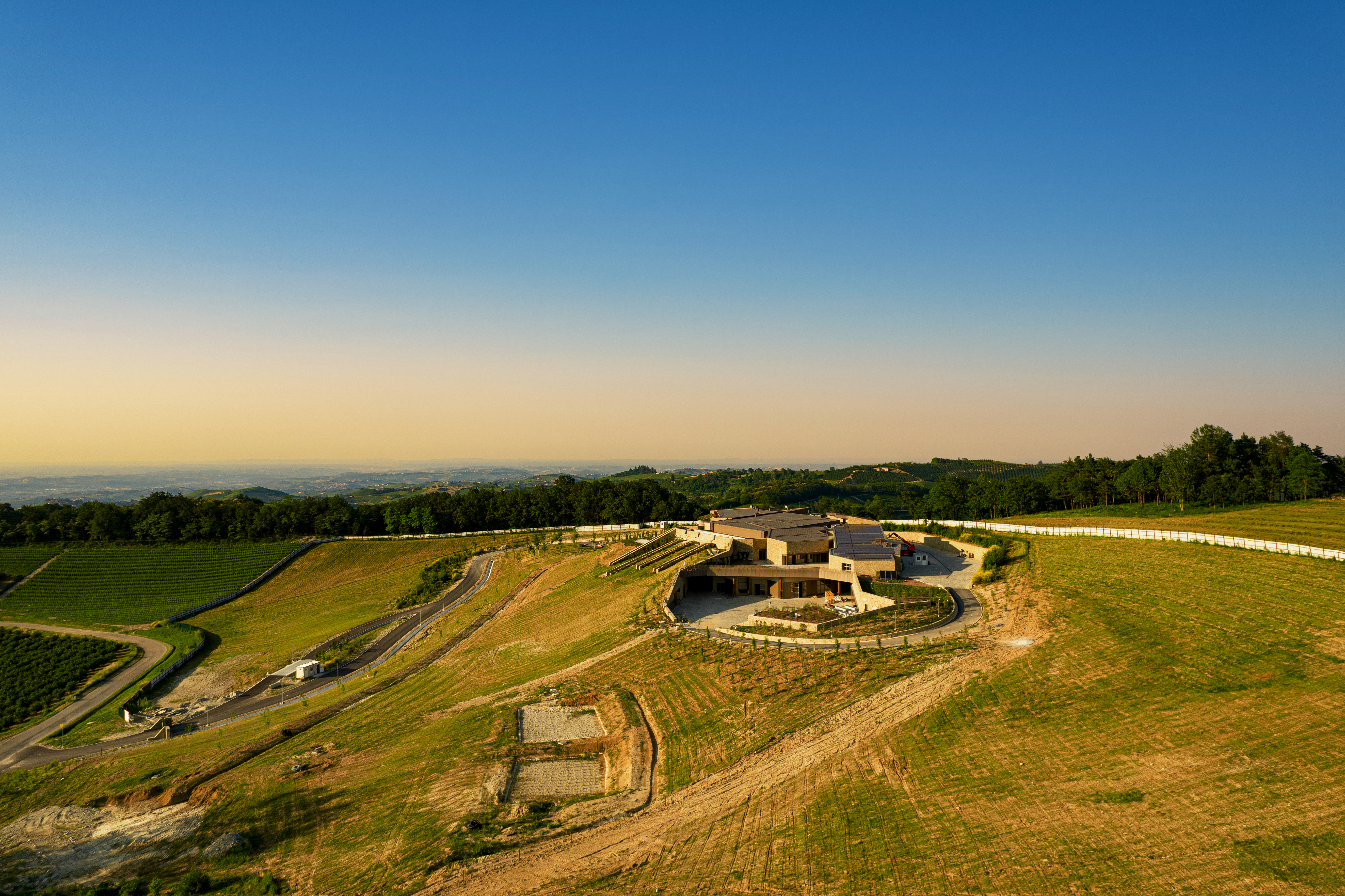 Eine moderne Weinbaustätte liegt malerisch auf einer Anhöhe, umgeben von sanften Hügeln und Weinreben. Der klare Himmel und die Landschaft schaffen eine ruhige Atmosphäre.