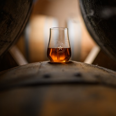 Ein Glas mit bernsteinfarbenem Getränk steht auf einem Holzfass. Im Hintergrund sind weitere Fässer sichtbar, die eine rustikale Atmosphäre schaffen.