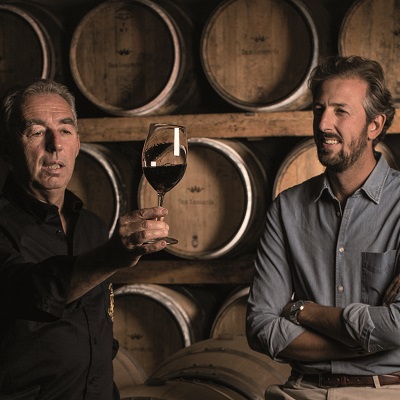 Zwei Männer stehen vor Holzfässern in einem Weinkeller. Einer hält ein Weinglas mit rotem Wein in die Höhe und spricht, während der andere aufmerksam zuhört.