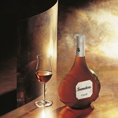 Eine Flasche Samalens VSOP Cognac steht neben einem eleganten Glas mit Getränk. Der Hintergrund zeigt sanfte, warme Lichtakzente und eine reflektierende Metalloberfläche.