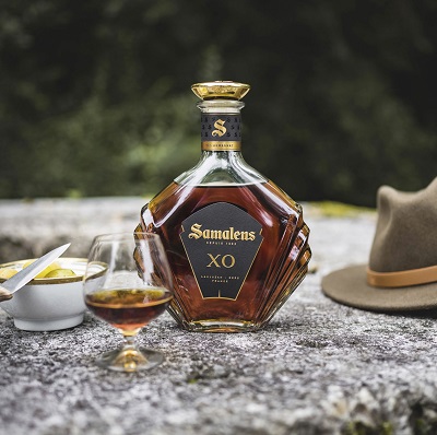 Eine elegante Flasche Samalens XO steht auf einem Stein, daneben ein Glas mit dem Getränk und ein kleiner Teller mit gelben Früchten. Ein brauner Hut liegt in der Nähe. Die Umgebung ist grün und naturbelassen.