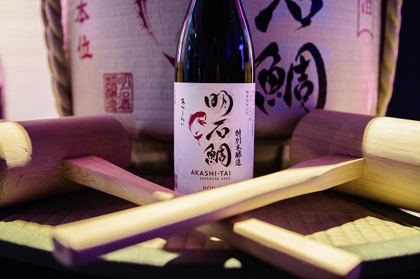 Eine Flasche Akashi-Tai Sake steht im Mittelpunkt, umgeben von zwei Holzstöcken und einem geflochtenen Untergrund. Im Hintergrund ist ein traditionelles japanisches Sake-Fass zu sehen.