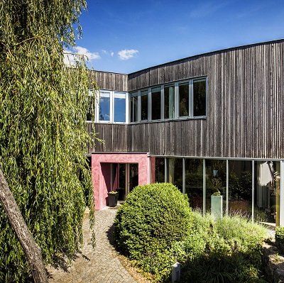 Ein modernes, zweistöckiges Haus aus Holz mit großen Fenstern. Im Vordergrund gibt es eine grüne Hecke und einen Gartenweg, umgeben von Bäumen und Büschen. Der Himmel ist klar und blau.