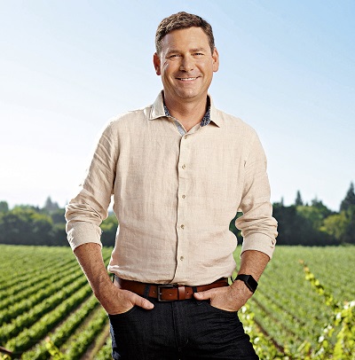 Ein Mann steht lächelnd mit beiden Händen in den Taschen vor einem Weinberg. Er trägt ein helles Hemd und Jeans, während der Himmel klar und blau ist. Im Hintergrund sind Weinstöcke sichtbar.