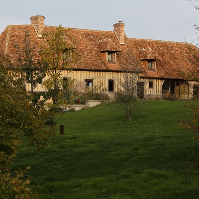 Ein traditionelles Landhaus mit einem roten Satteldach. Das Gebäude besteht aus Holz, hat mehrere Fenster und befindet sich auf einer grünen Wiese, umgeben von Bäumen und Pflanzen.