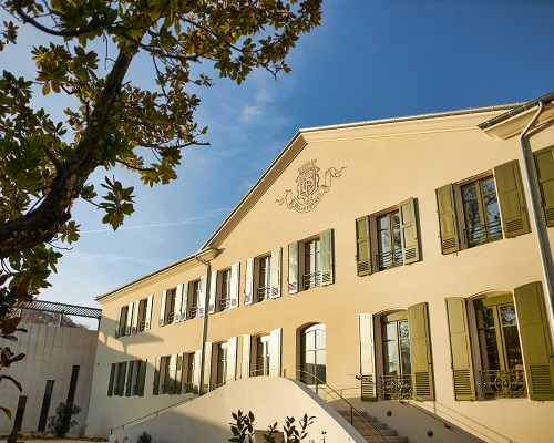 Ein historisches Gebäude mit einem hellen Putz und grünen Fensterläden. Die Fassade hat ein Wappen und ist von Bäumen umgeben. Der Himmel ist blau mit wenigen Wolken.