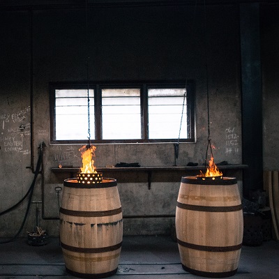 Zwei Holzfässer stehen in einer dunklen Werkstatt, aus denen Flammen aufsteigen. Im Hintergrund gibt es ein Fenster und eine Arbeitsfläche mit Werkzeugen. An den Wänden sind Handschriftliche Notizen zu sehen.