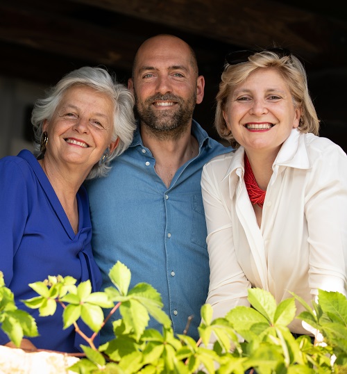 Drei Personen lächeln freundlich und stehen zusammen vor einer rustikalen Kulisse. Eine Frau trägt ein blaues Oberteil, die andere eine weiße Bluse, der Mann ein blaues Hemd. Grüne Pflanzen umgeben sie.