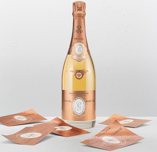 Eine Flasche Louis Roederer Cristal Rosé 2020 steht auf einem hellen Hintergrund. Rundherum sind mehrere orangefarbene Etiketten verteilt, die das Logo der Marke zeigen.