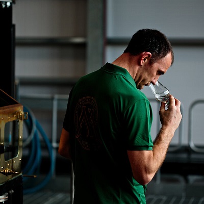 Ein Mann in einem grünen Poloshirt hält ein Glas in der Hand und riecht daran. Er steht in einer industriellen Umgebung mit metallischen Elementen und Rohren im Hintergrund.
