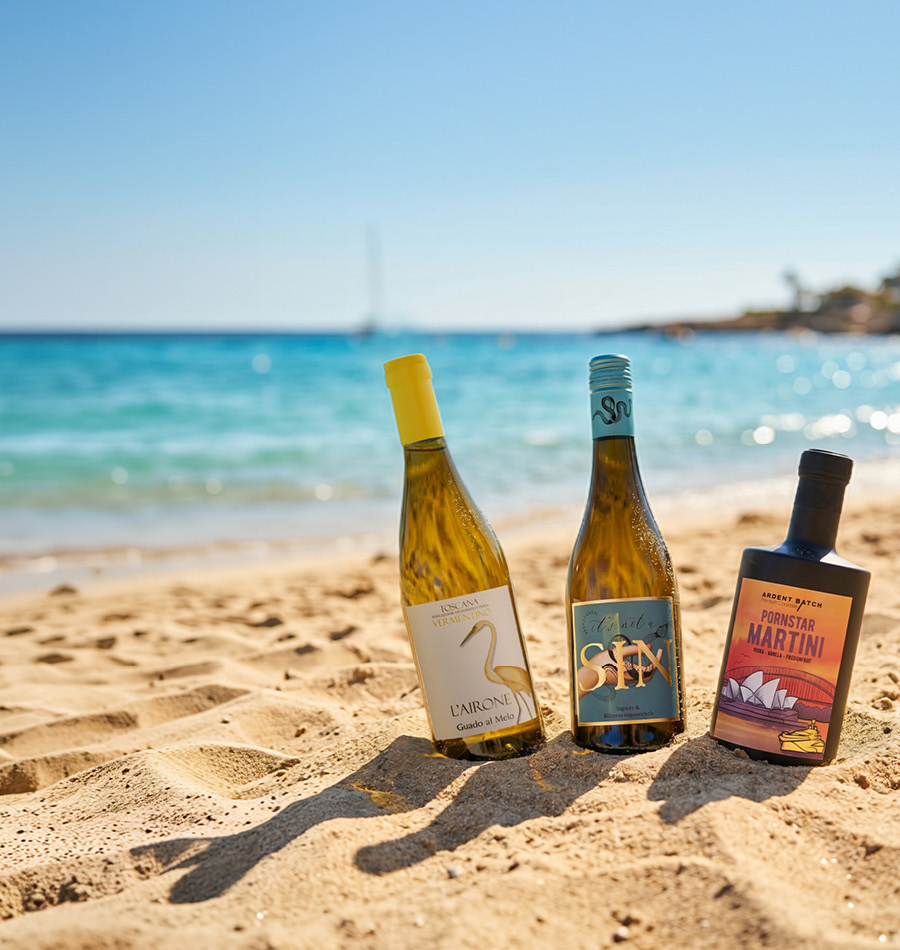Drei Flaschen Wein stehen auf sandigem Strandboden. Im Hintergrund glitzert das blaue Wasser des Meeres unter klarem, sonnigem Himmel. Ein entspannender Urlaubsatmosphäre wird vermittelt.