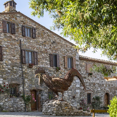 Ein großes, rustikales Steinhaus mit mehreren Fenstern und Holzläden. Vor dem Haus steht eine große, skulpturale Hahnstatue auf einem kleinen Steinpodest. Umgeben von Bäumen und Pflanzen.