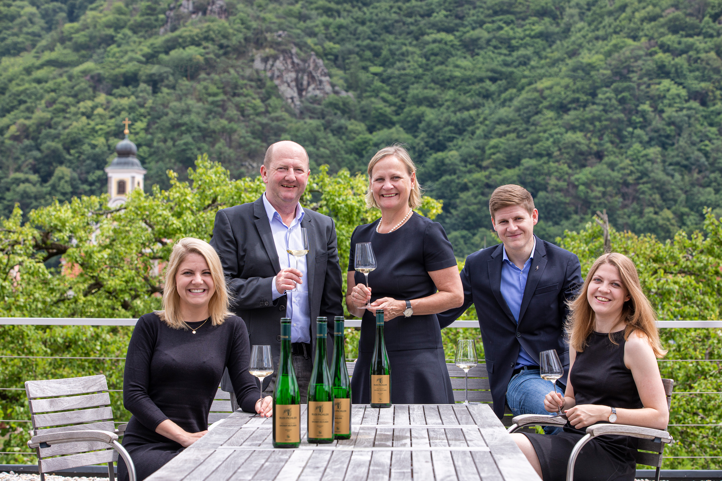 Eine Gruppe von fünf Personen steht und sitzt an einem Holztisch, umgeben von einer grünen Berglandschaft. Sie halten Weingläser und präsentieren mehrere Flaschen Wein, während sie lächeln.