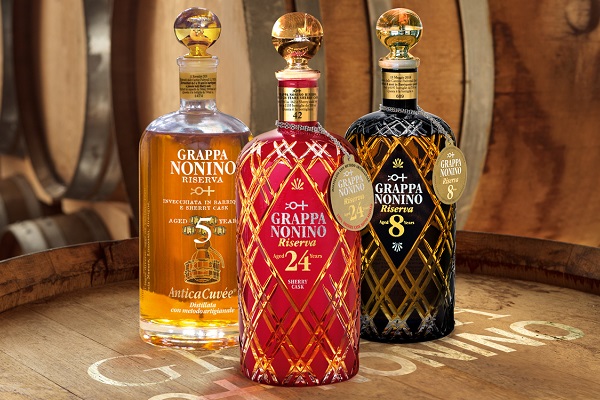 Drei Flaschen Grappa stehen auf einem Holzfass. Die Flaschen sind unterschiedlich farbig: eine goldene, eine rote und eine schwarze mit dekorativen Etiketten. Im Hintergrund sind Holzfässer sichtbar.
