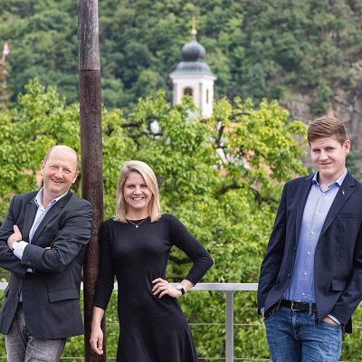 Drei Personen stehen entspannt auf einer Terrasse mit Blick auf grüne Bäume und eine Kirche im Hintergrund. Die Männer tragen Anzüge, die Frau ein schwarzes Kleid.