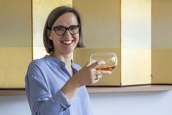 Eine Frau mit Brille lächelt und hält ein Glas mit einem Getränk in der Hand. Im Hintergrund sind goldene Paneele zu sehen. Sie trägt ein hellblaues Hemd.