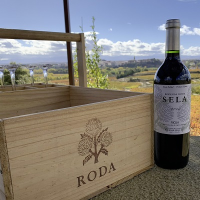 Ein Holzkorb mit dem Schriftzug -RODA- steht auf einer Steinfläche. Daneben befindet sich eine Flasche roten Weins mit dem Etikett -Bodega Roda Sela-. Im Hintergrund erstreckt sich eine malerische Landschaft mit Weinbergen und einem bewölkten Himmel.