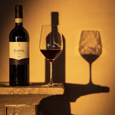 Eine Weinflasche mit dem Etikett -Bindella- steht neben einem Weinglas, das roten Wein enthält. Der Schatten des Glases und der Flasche fällt auf eine helle Wand.