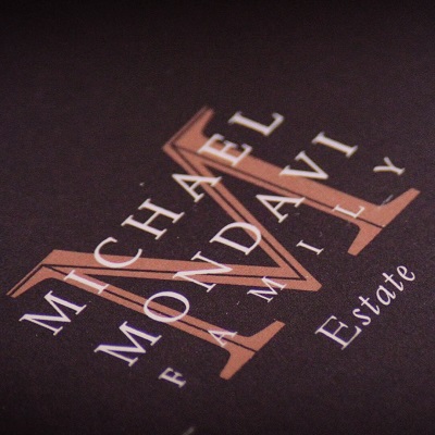 Ein Logo zeigt den Schriftzug -MICHAEL MONDAVI FAMILY Estate- in verschiedenen Schriftarten und Größen auf einem schwarzen Hintergrund. Die Buchstaben sind in Brauntönen gehalten.