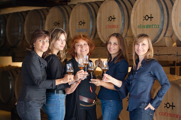 Fünf Frauen stehen in einem Weinkeller, halten Gläser mit Wein und lächeln. Im Hintergrund sind Holzfässer zu sehen, auf denen bunte Namen stehen. Die Atmosphäre ist festlich und freundlich.