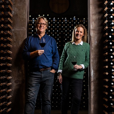 Zwei Personen stehen in einem Weinkeller, jede mit einem Glas Rotwein in der Hand. Die Wand im Hintergrund ist mit Weinflaschen gefüllt. Die Umgebung ist dunkel und gemütlich.
