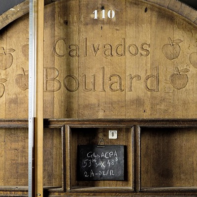 Ein großes Holzfass mit der eingravierten Aufschrift -Calvados Boulard- und Äpfeln. Ein kleines schwarzes Schild darunter zeigt Maße und Nummern an. Die Umgebung wirkt rustikal und traditionell.