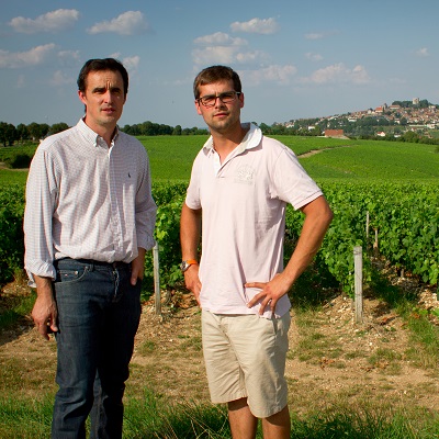 Zwei Männer stehen in einem Weinberg. Der Linke trägt ein kariertes Hemd und der Rechte ein weißes T-Shirt. Im Hintergrund sind Weinreben und eine Stadtansicht zu sehen, unter blauem Himmel mit einigen Wolken.