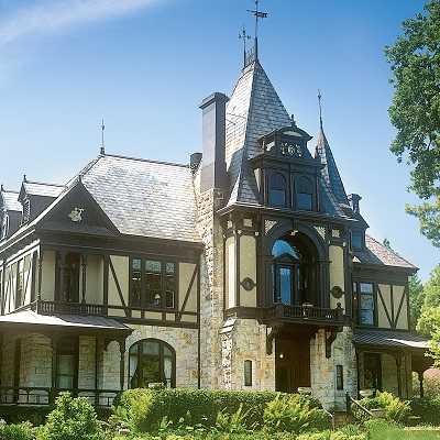 Ein großes, elegantes Haus im viktorianischen Stil mit einer Mischung aus Stein- und Holzelementen. Es hat spitze Dächer, große Fenster und eine verzierten Fassade, umgeben von üppigem Grün.