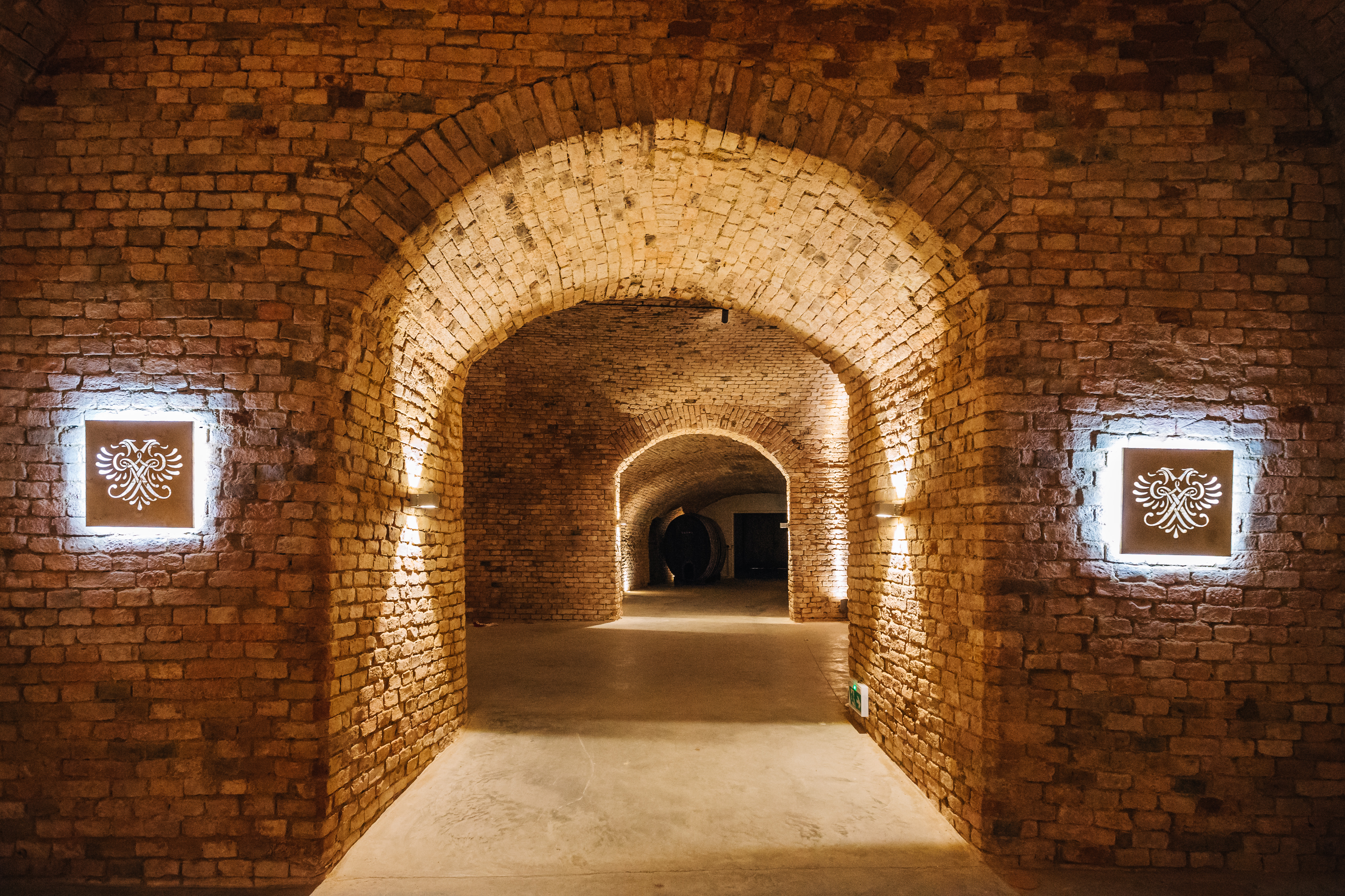 Ein gewölbter Raum aus Ziegelsteinen mit beleuchteten Wandschildern und weichem Licht, das die Wände erhellt. Im Hintergrund sind weitere Bögen und ein Holzfass zu sehen.