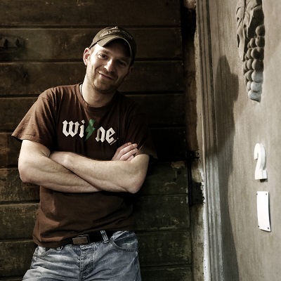 Ein Mann mit einem braunen T-Shirt und Bluejeans lehnt entspannt an einer Wand aus Holz. Er lächelt und hat eine Kappe auf, während eine Tür mit der Nummer 2 im Hintergrund sichtbar ist.