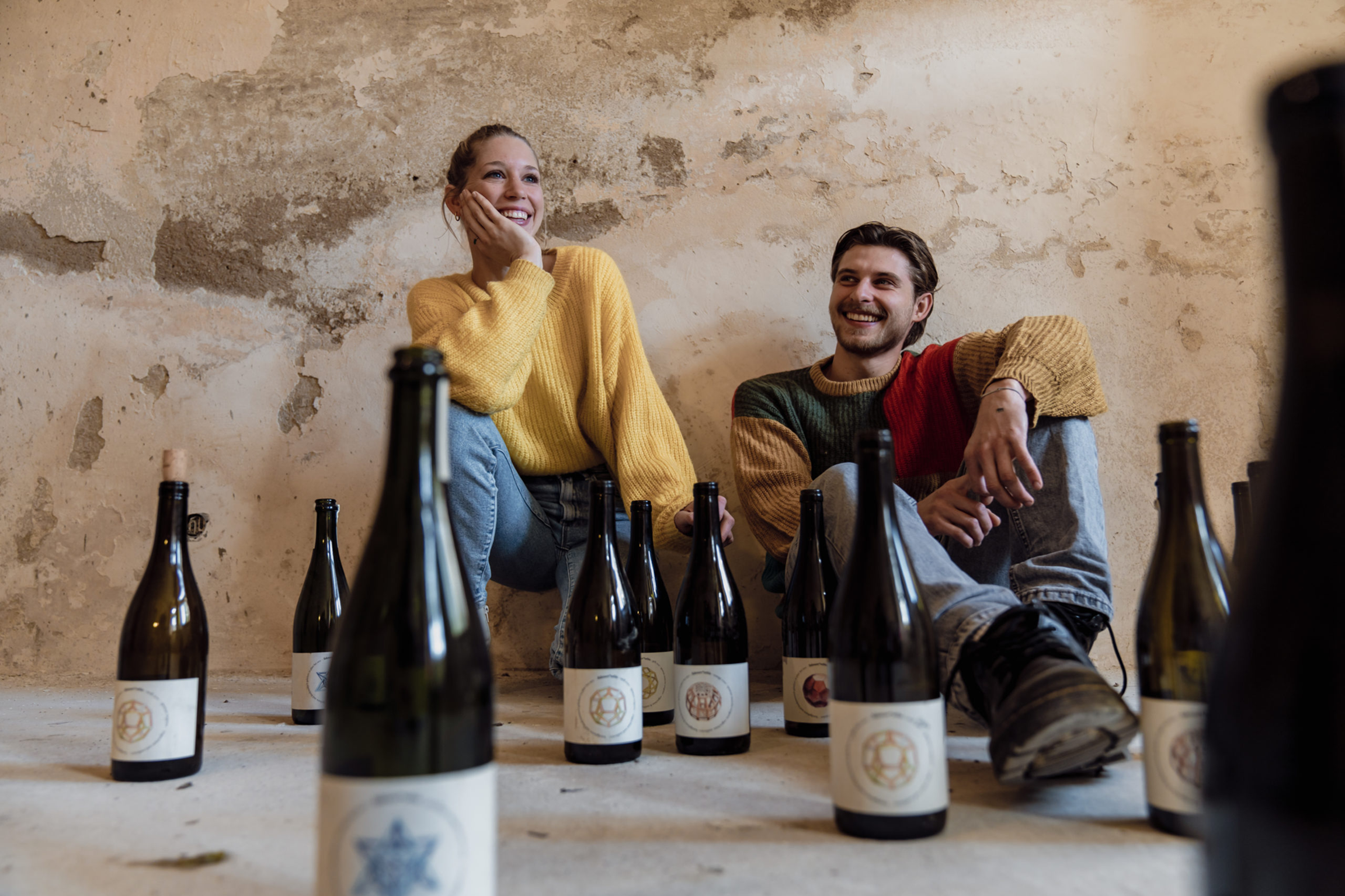 Zwei Personen sitzen fröhlich am Boden, umgeben von Weinflaschen. Die Wände sind uneben und zeigen eine graue Patina. Die Atmosphäre ist entspannt und freundlich.