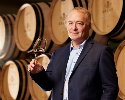 Ein Mann in einem Anzug hält ein Weinglas mit Rotwein in der Hand. Im Hintergrund sind große Weinfässer angeordnet. Die Atmosphäre wirkt elegant und professionell.